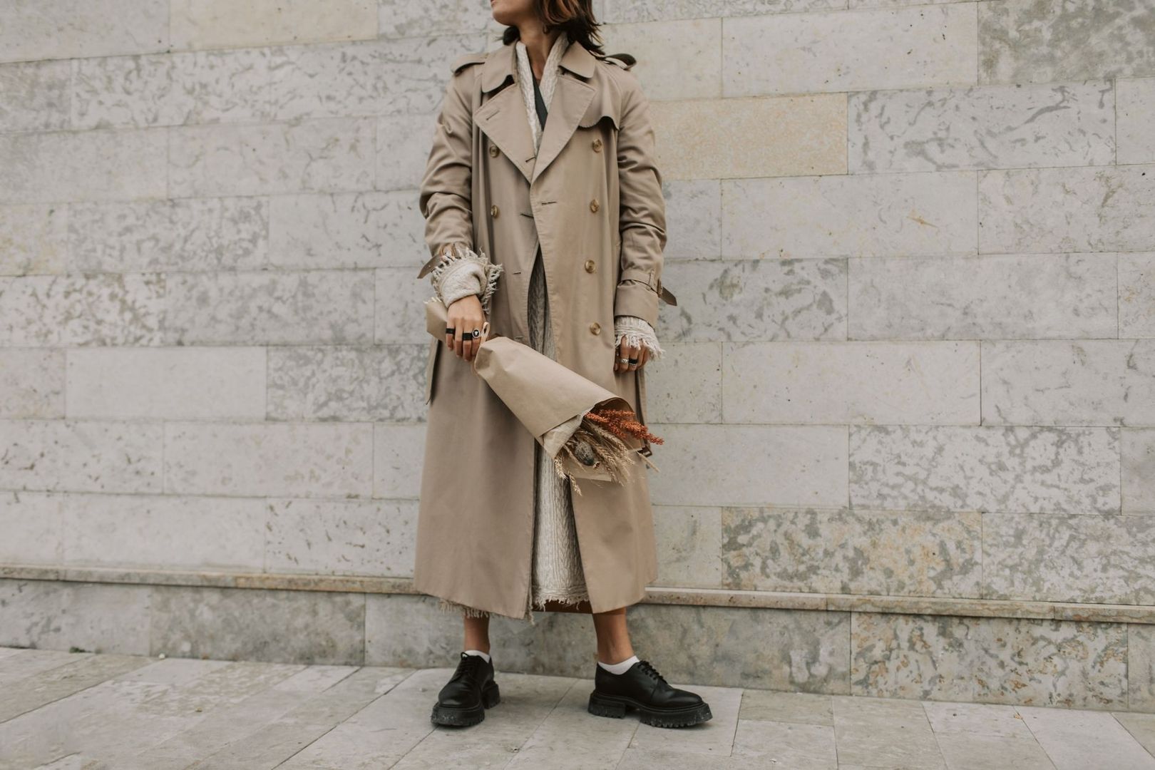 Person i beige trenchcoat med bukett framför en stenmur